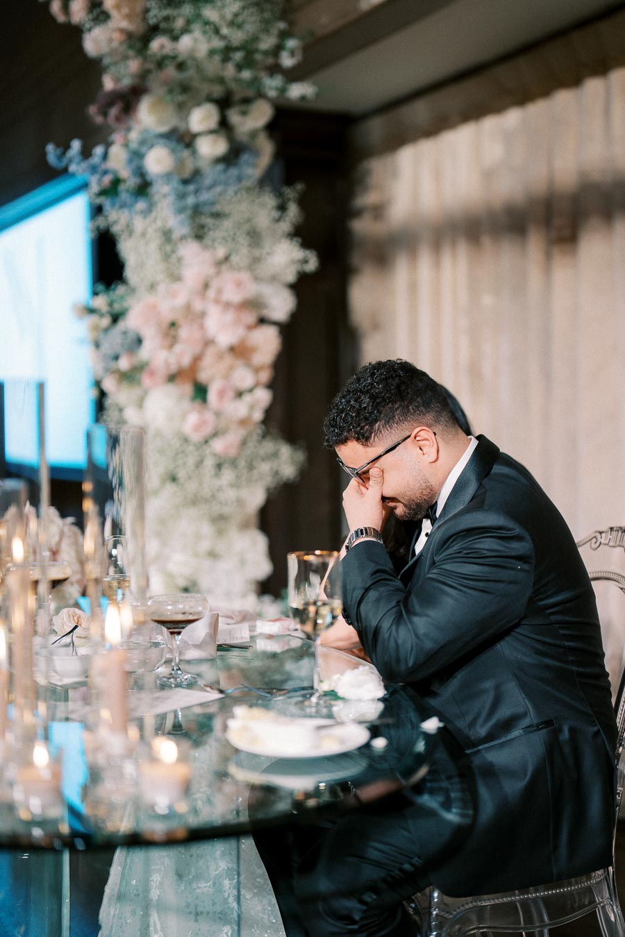 A man in a suit sitting at an elegantly set wedding reception table, appearing emotional with his hand covering his face, surrounded by candles and floral arrangements.