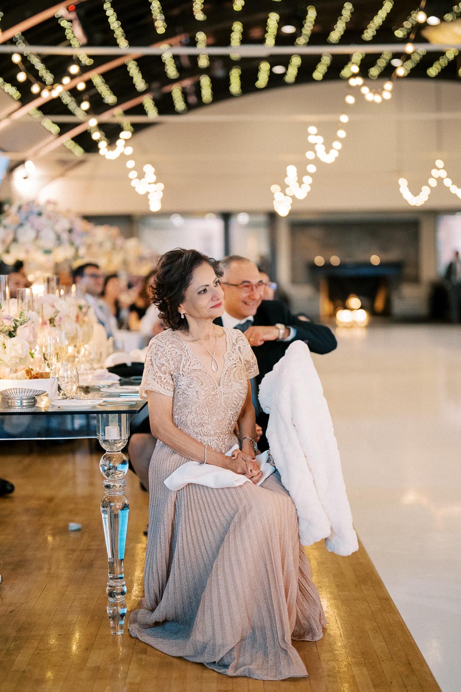 Elegant woman in a lace dress sitting at a beautifully decorated wedding reception, surrounded by soft candlelight and floral arrangements, with guests and warm lighting in the background.