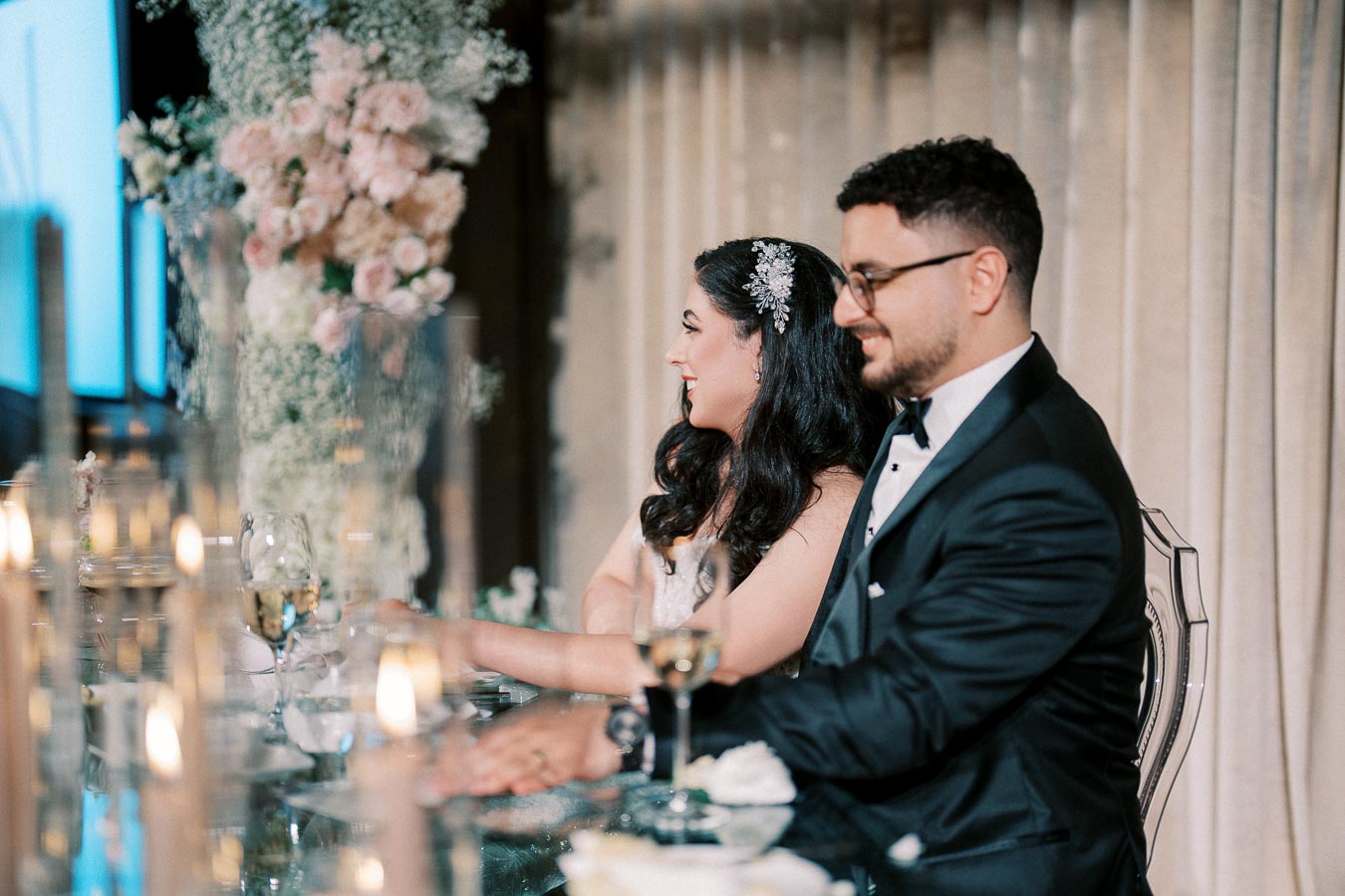 Elegant wedding reception scene with a smiling couple seated at a beautifully decorated table, surrounded by delicate floral arrangements and glowing candlelights, during a joyful celebration.