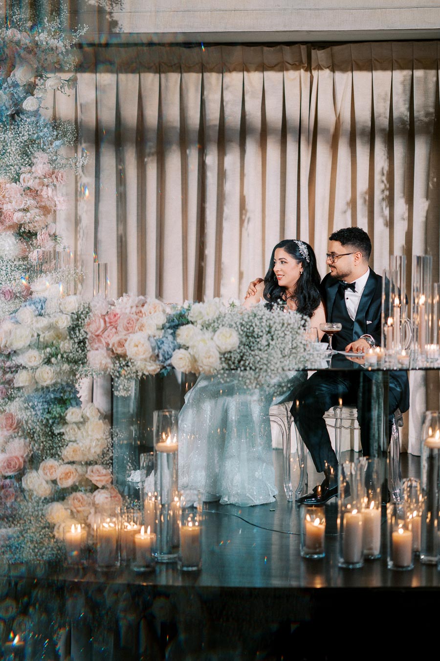 A bride and groom sitting together at a wedding reception table adorned with elegant floral arrangements and lit candles, creating a romantic and luxurious ambiance.