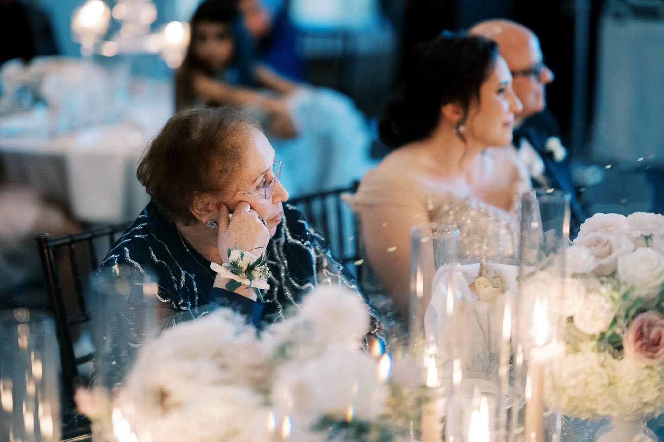 Elderly woman at a wedding reception, gazing thoughtfully, surrounded by elegant floral arrangements and soft candlelight.