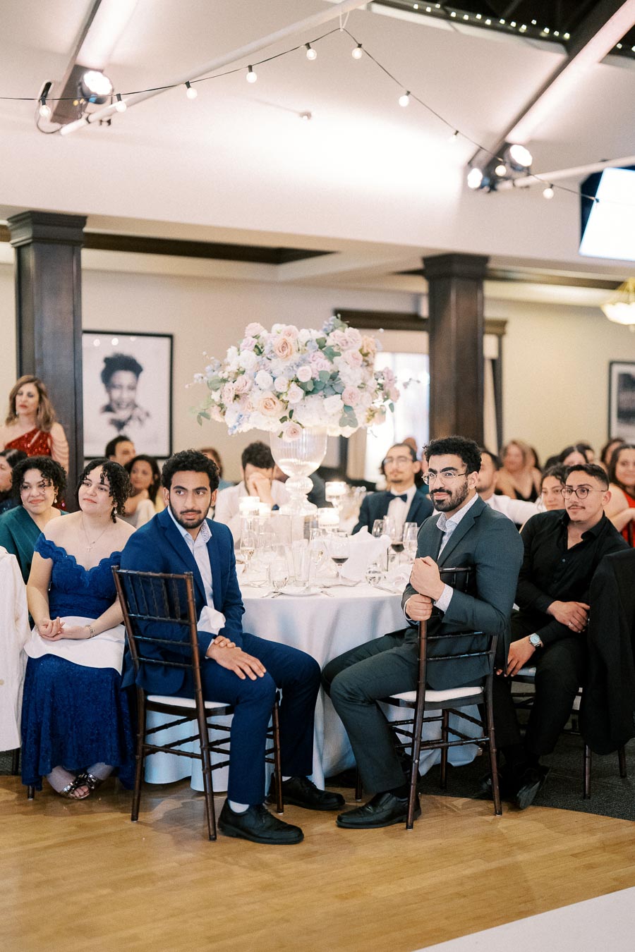 Guests seated around an elegantly decorated table at a formal event, featuring a floral centerpiece and ambient lighting, capturing a moment of anticipation and interaction.