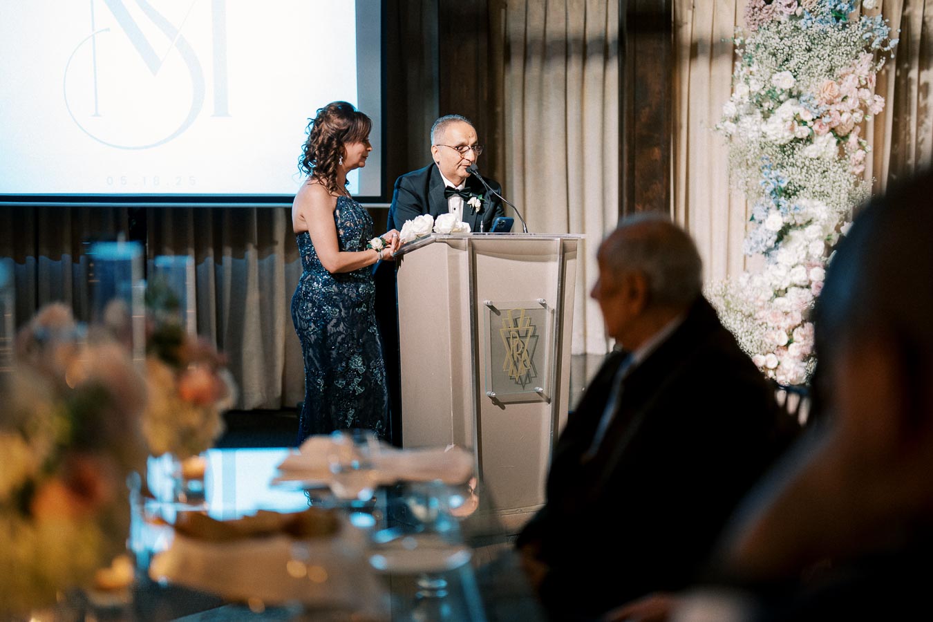 A couple elegantly dressed, standing at a podium during a formal event, with floral decorations and a projected monogram in the background, creating a sophisticated ambiance.