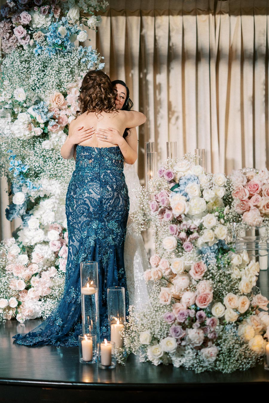 A heartfelt embrace between two women at a wedding ceremony, surrounded by lush floral arrangements with pink, white, and blue flowers. Candles in glass holders add a warm glow to the elegant scene.