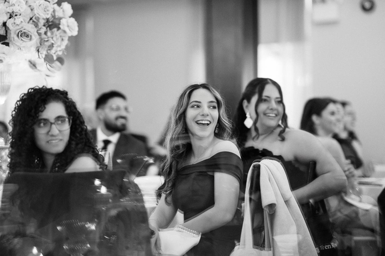 A group of people smiling at a formal event, elegantly dressed, sitting at a table adorned with floral decorations. Black and white photo capturing a joyful moment.