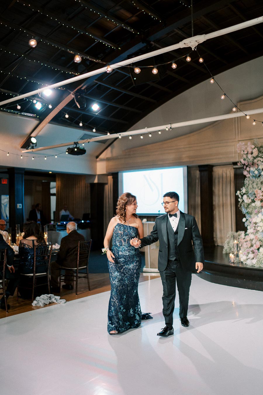 A couple elegantly dressed in formal attire dances on a lit-up floor at a beautifully decorated wedding reception venue, with guests seated at tables and soft lighting creating an intimate ambiance.