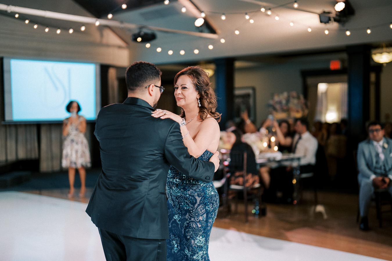 A couple elegantly dances together during a formal event, surrounded by guests seated at decorated tables under string lights. The woman wears a sparkling blue gown, while the man is dressed in a black suit.