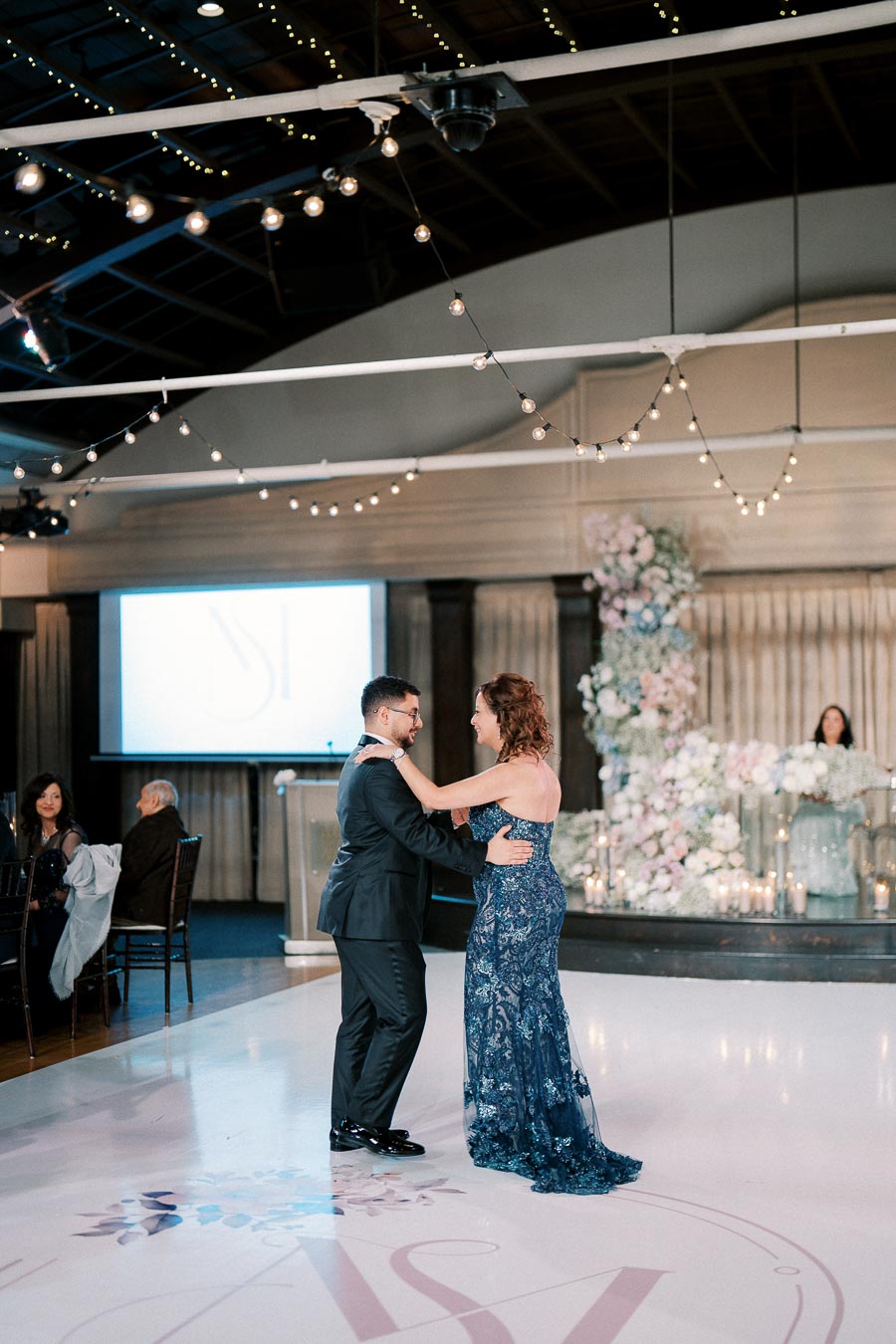 A couple shares a romantic dance on a decorated white dance floor at an elegant wedding reception, surrounded by string lights and floral arrangements, with seated guests and a large display screen in the background.