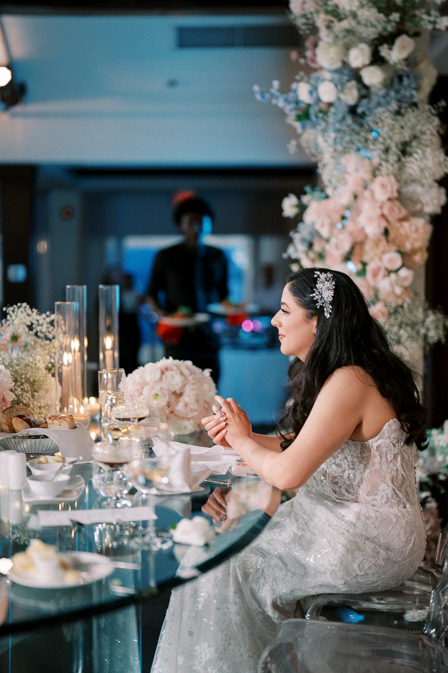 A bride in a lace wedding gown sits at a glass table during a wedding reception, with elegant floral arrangements and lit candles in the background.