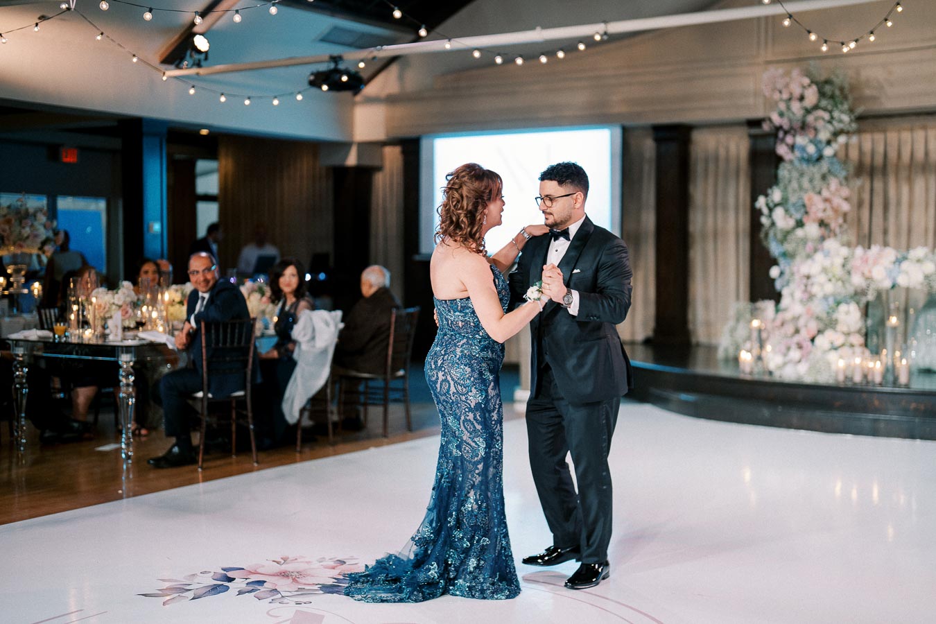 A couple elegantly dancing at a wedding reception, the woman in a blue lace gown and the man in a black tuxedo, surrounded by seated guests and floral decorations under string lights.