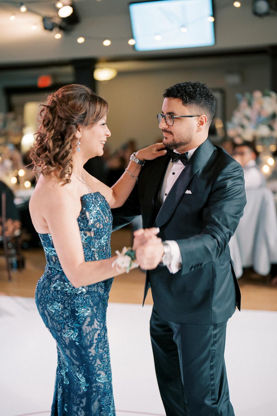 A couple gracefully dancing at a formal event, with the woman wearing an elegant blue gown and the man in a classic black tuxedo, surrounded by softly lit decor and romantic ambiance.