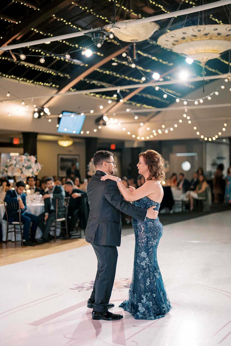 A couple elegantly dancing at a wedding reception under twinkling string lights with guests seated at tables in the background.