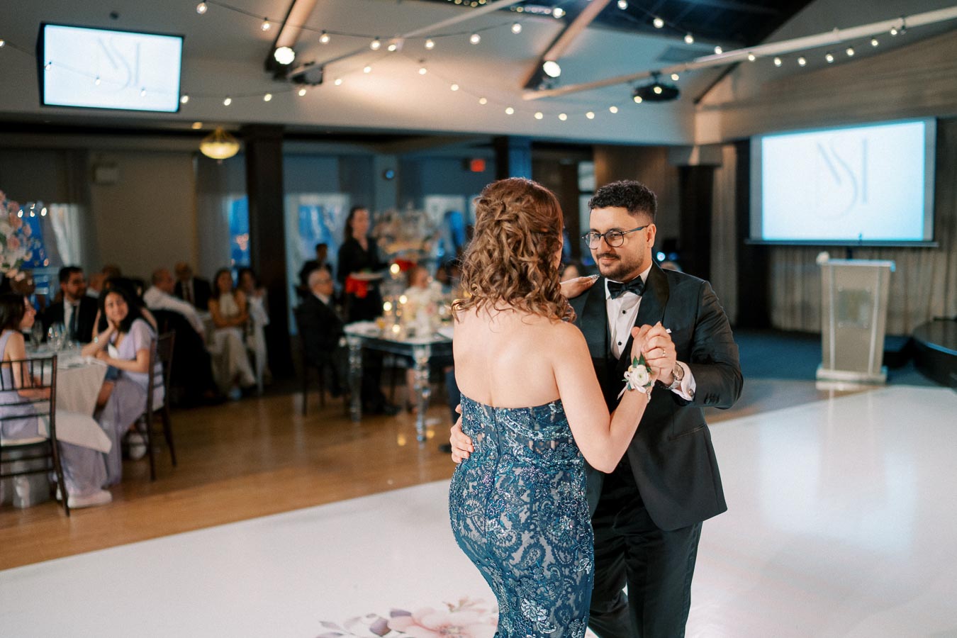 A couple elegantly dancing in formal attire at a beautifully decorated indoor wedding reception, with guests seated at tables in the background and string lights creating a warm ambiance.