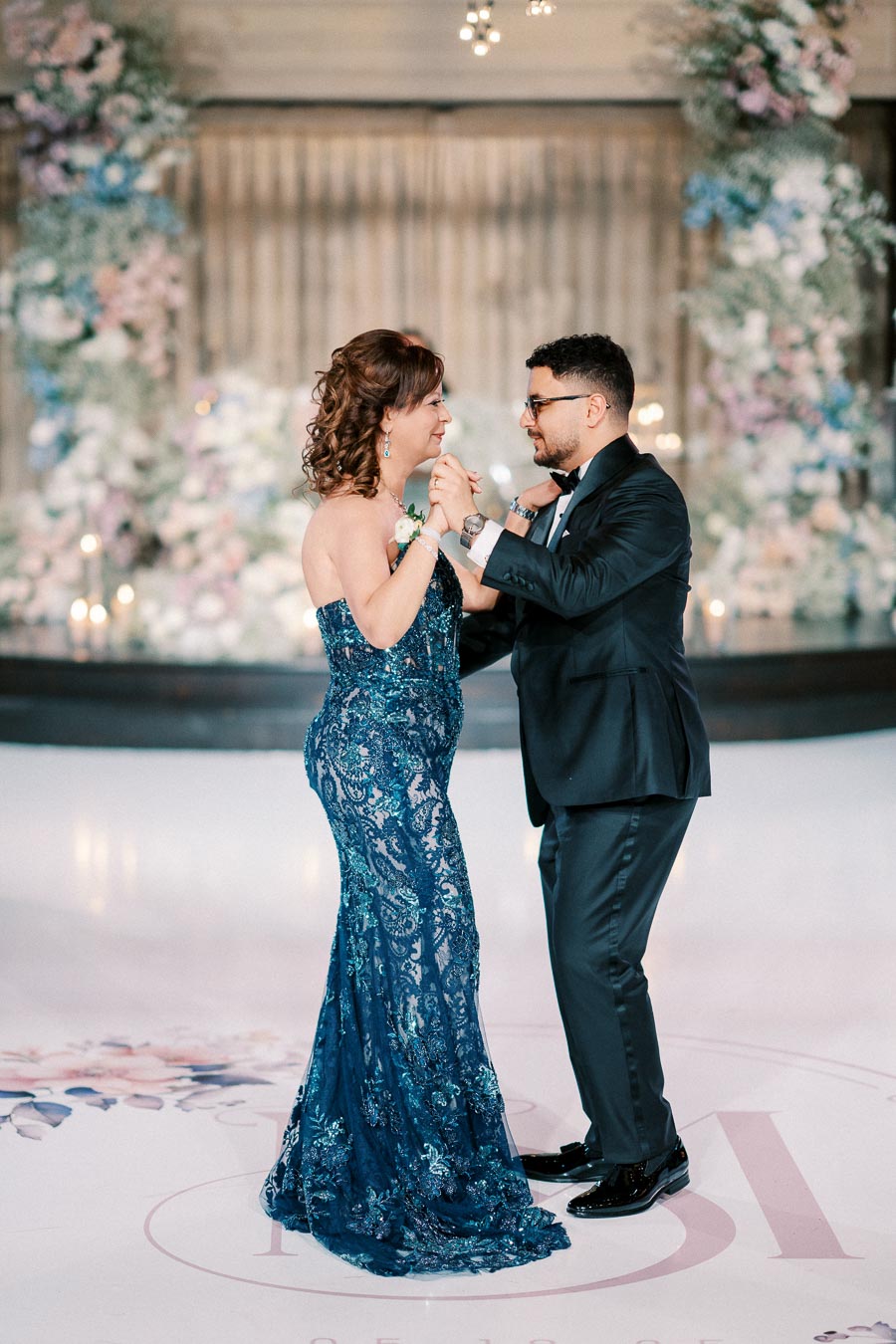 A couple elegantly dances at a wedding, with the woman in a blue lace gown and the man in a black tuxedo, against a backdrop of floral decorations.