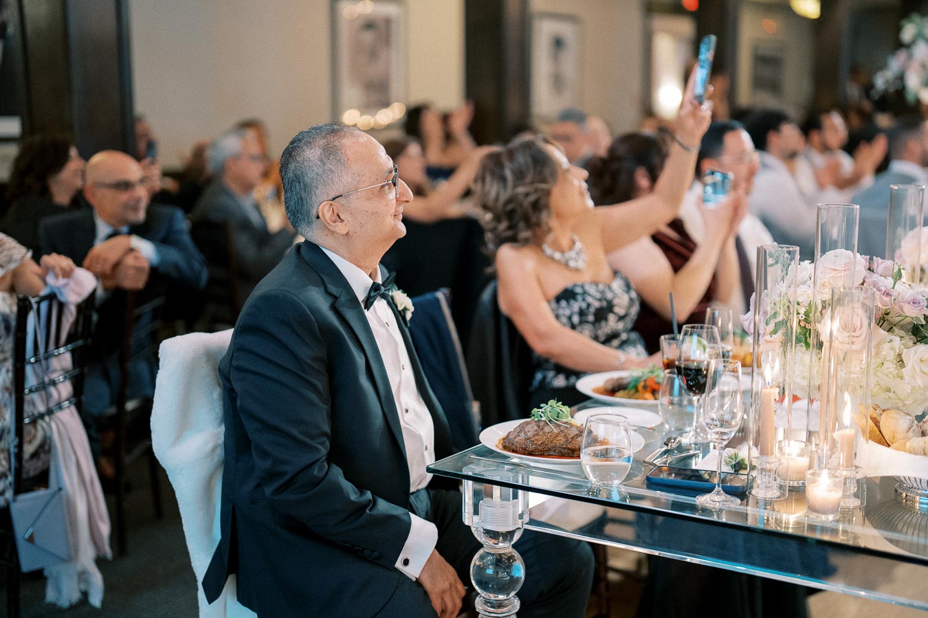 Elderly gentleman in a tuxedo seated at an elegantly set dinner table, surrounded by guests capturing moments on their phones at a formal event, with floral arrangements and lit candles enhancing the ambiance.