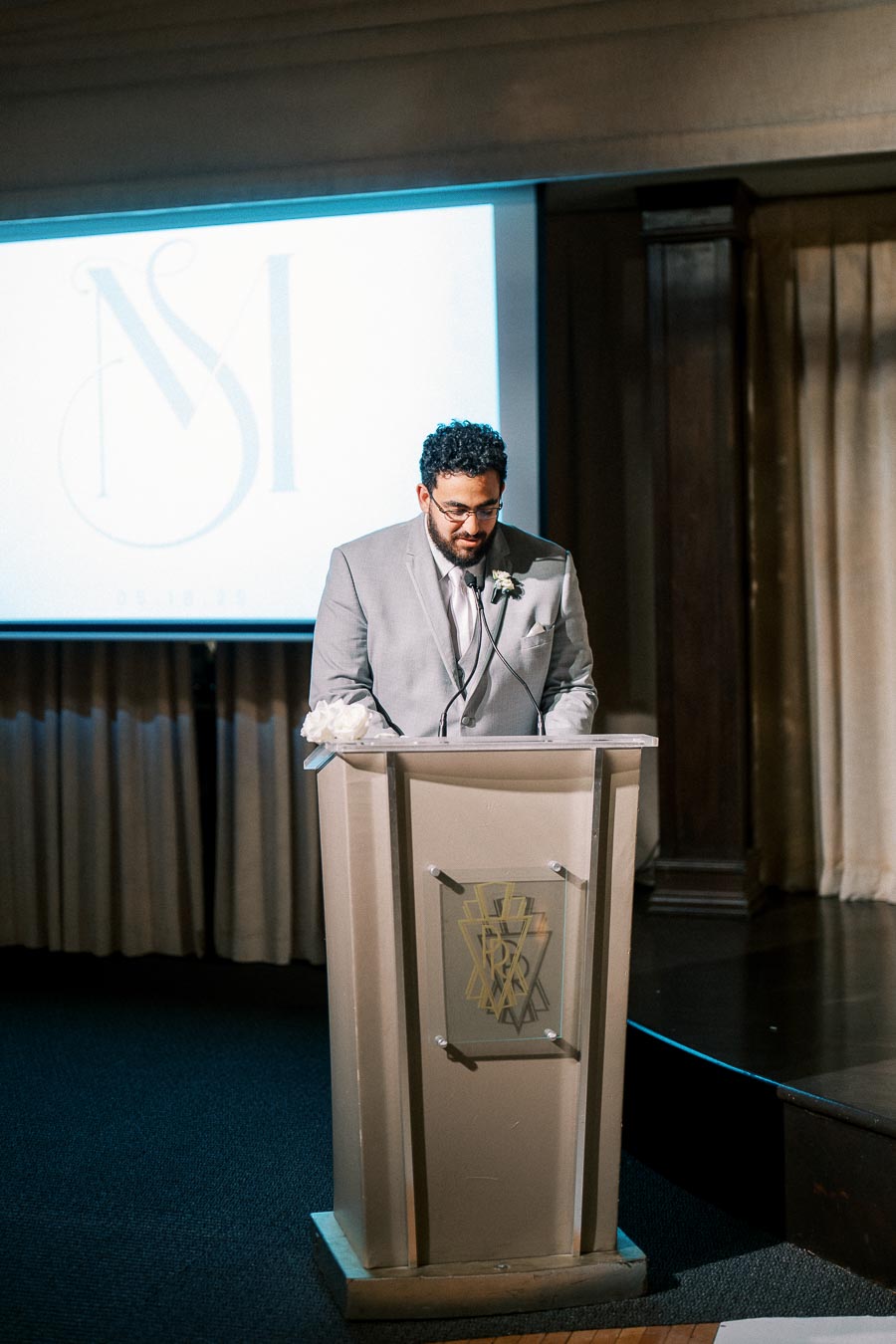 Man in a gray suit giving a speech at a podium during an indoor event, with a screen displaying a logo in the background.