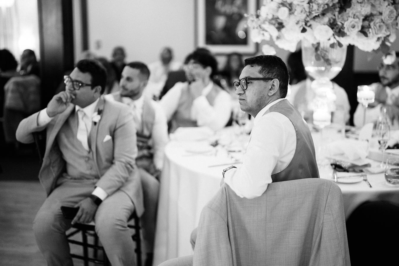A black and white photo of well-dressed wedding guests seated around a table, attentively listening during a wedding reception, with elegant floral arrangements in the background.
