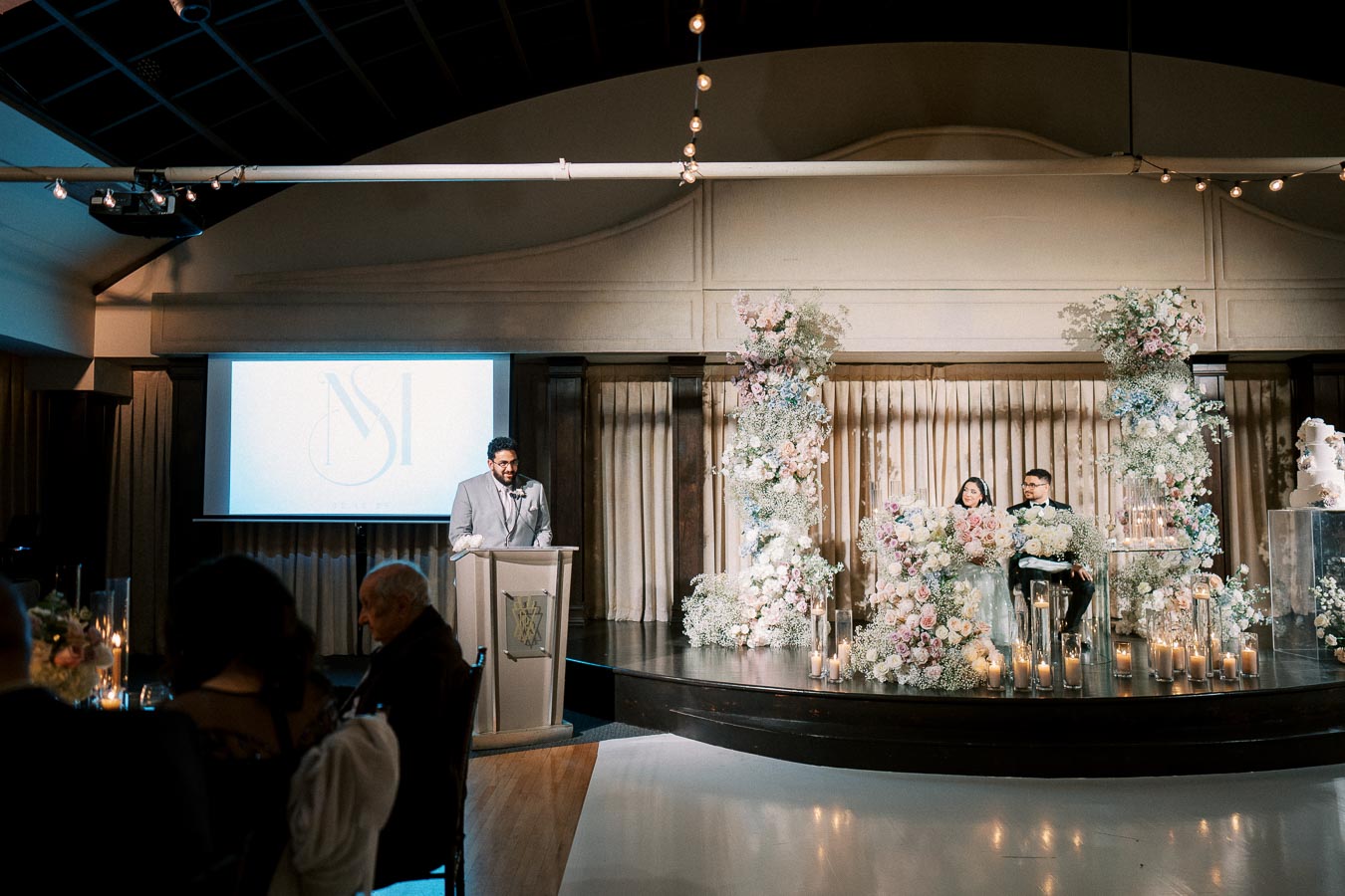 Elegant wedding reception hall featuring a speaker at a podium, beautifully decorated stage with lush floral arrangements and candles, and a screen displaying a logo.