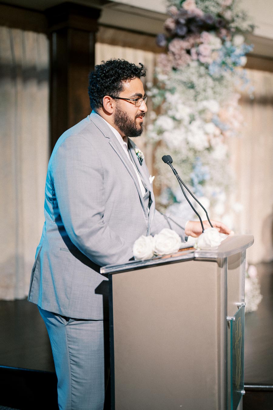 A man in a light gray suit speaking at a podium, decorated with white roses, during an event in a floral-themed venue.