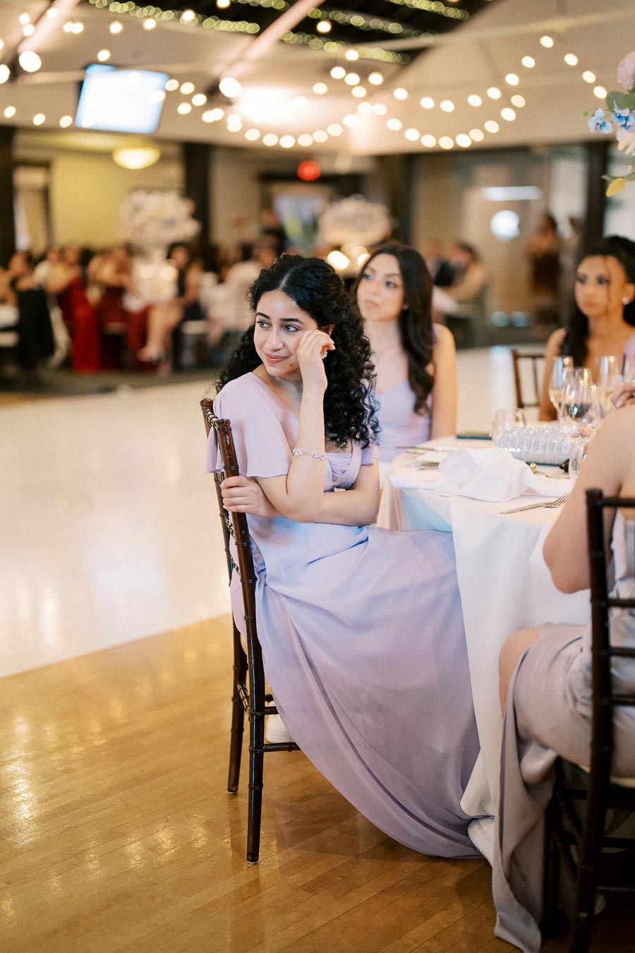 A woman in a lavender dress sits at a round table during a formal event, surrounded by softly lit string lights, in a warmly decorated banquet hall. Guests are visible in the background.