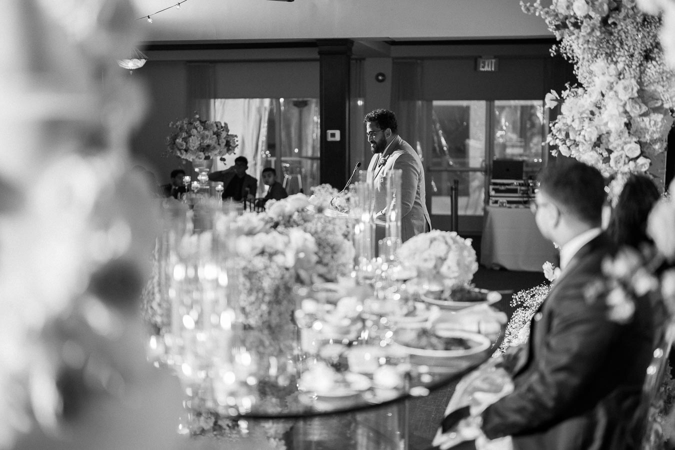 Black and white image of a wedding reception, featuring a man in a suit giving a speech at a decorated table, with floral arrangements and guests in the background.