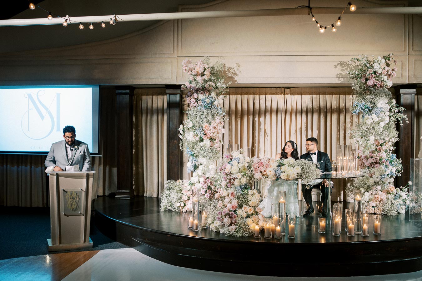 A couple sits at a beautifully decorated sweetheart table with pastel floral arrangements and candles, while a man in a gray suit delivers a speech at a podium during a wedding reception.