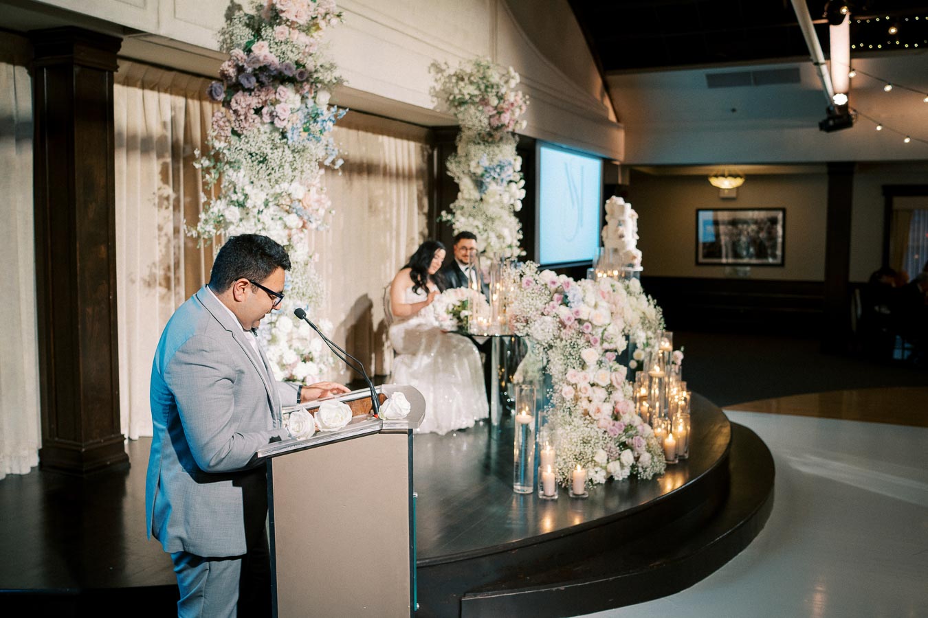 Man in a gray suit giving a speech at a wedding reception, with the bride and groom seated at a decorated table adorned with flowers and candles in the background.