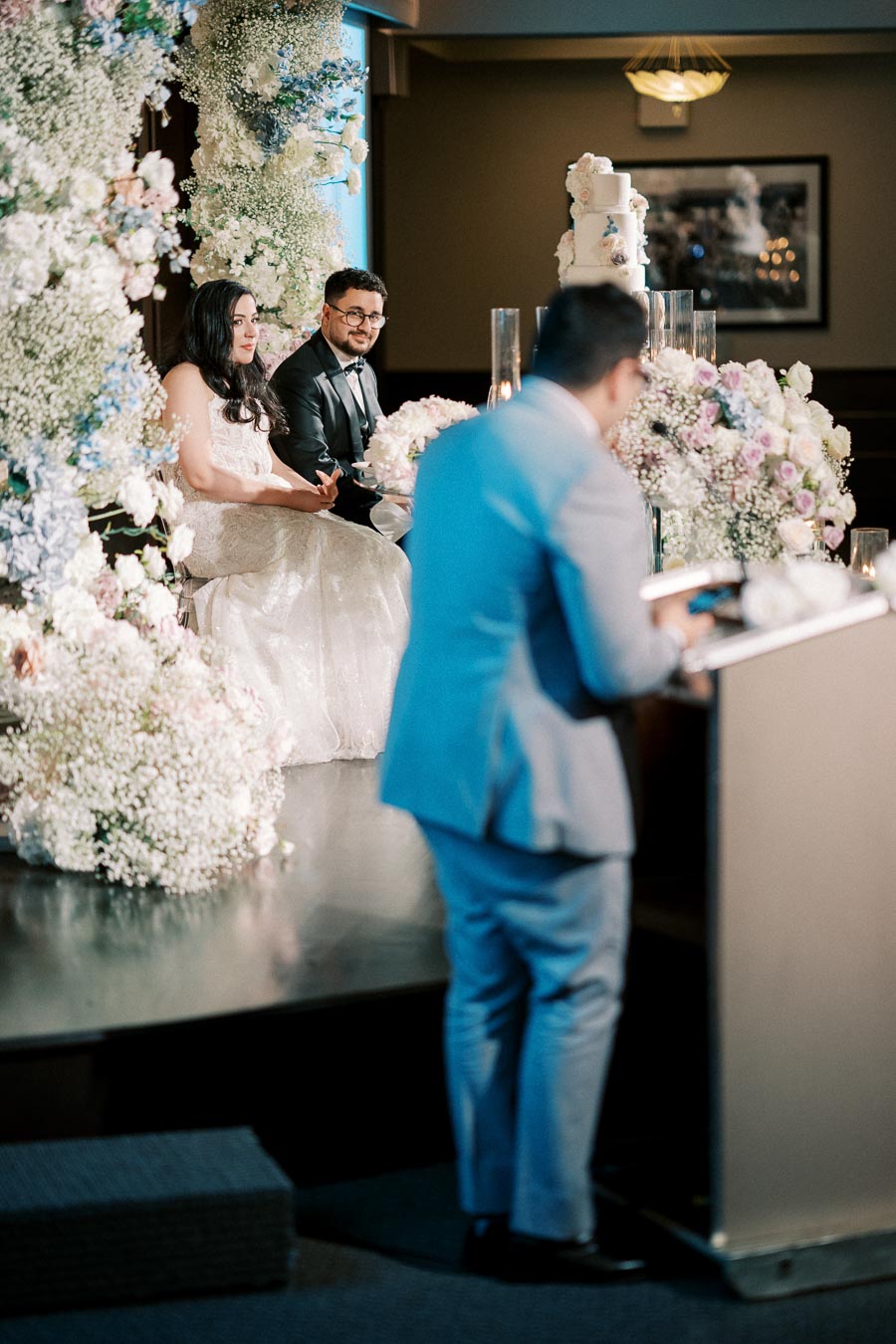 Bride and groom seated at a flower-adorned reception table, listening to a speech by a groomsman in a blue suit, with a multi-tiered wedding cake in the background.