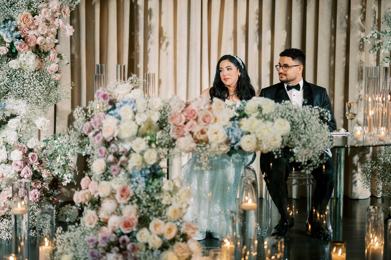 Elegant wedding reception with a couple sitting at a lavishly decorated table, surrounded by pastel floral arrangements and glimmering candles, in a luxurious setting.