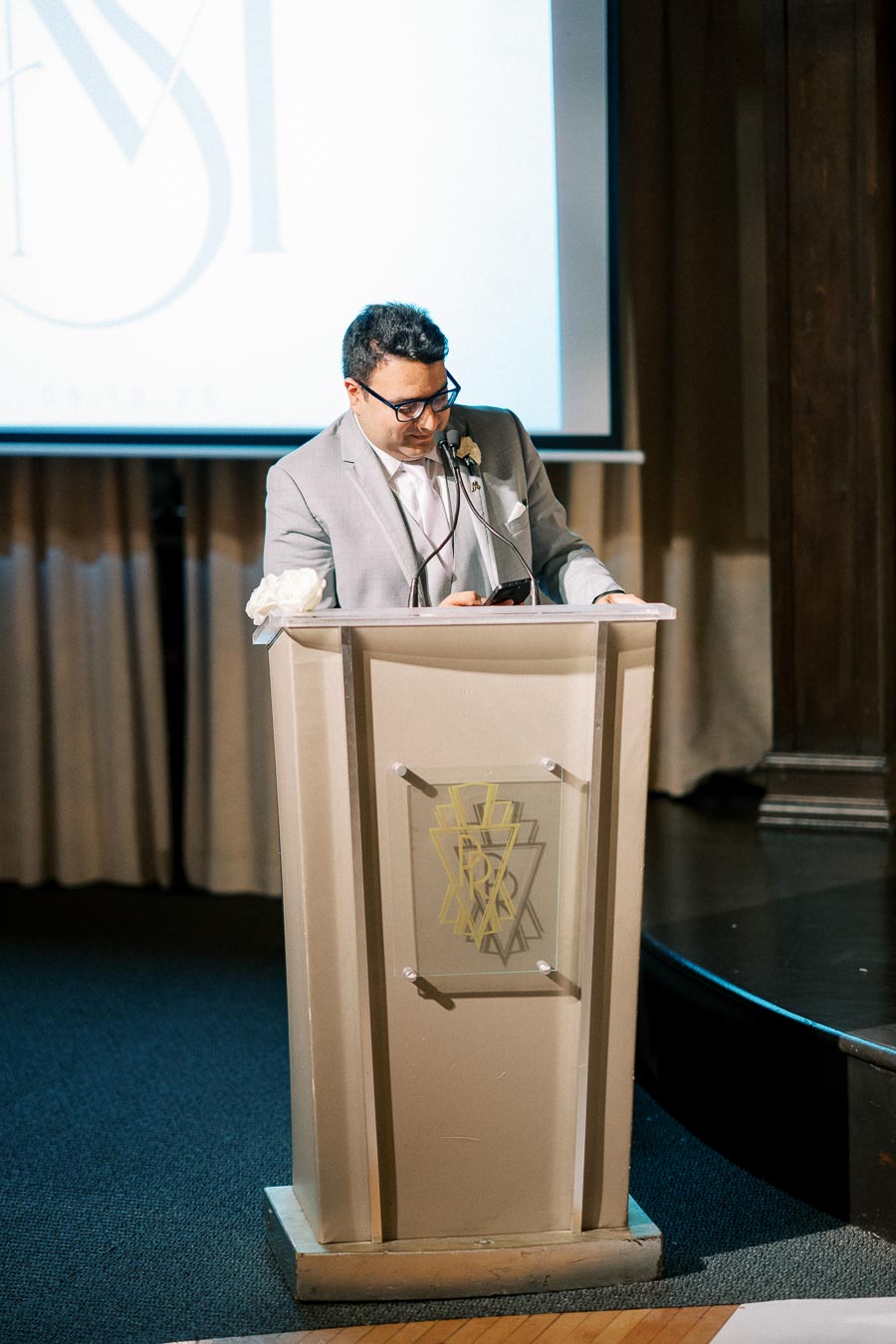 Man in a gray suit giving a speech at a podium with a microphone, in front of a screen, during an indoor event.