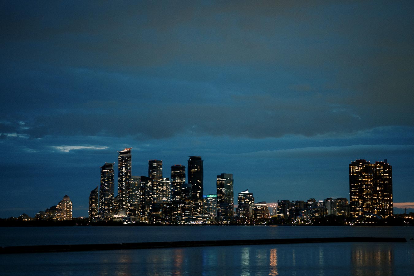 Alt text: Night view of a city skyline with illuminated skyscrapers reflected on the water, under a dark, cloudy sky.