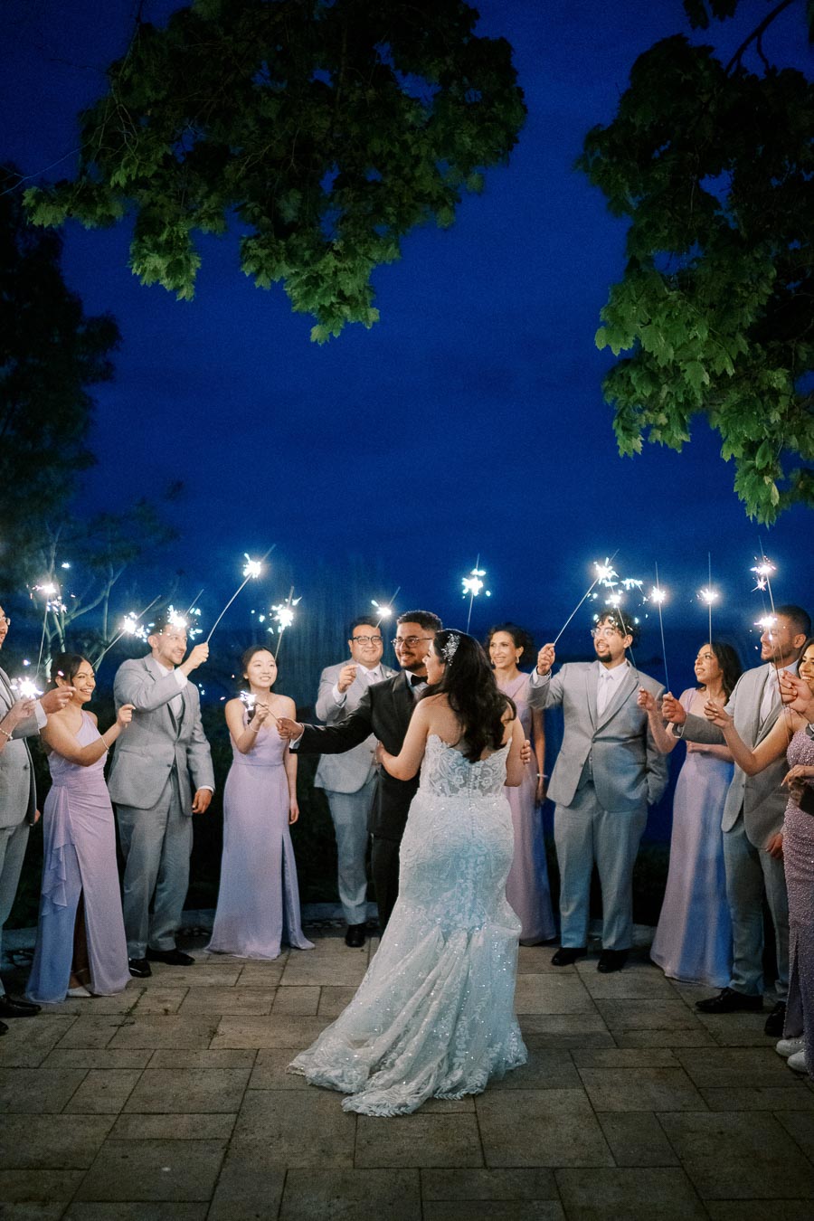 Bride and groom enjoying their first dance surrounded by wedding party holding sparklers, evening celebration under a clear blue sky.