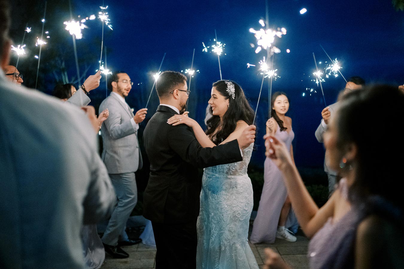A newlywed couple dances surrounded by guests holding sparklers during an evening wedding celebration.