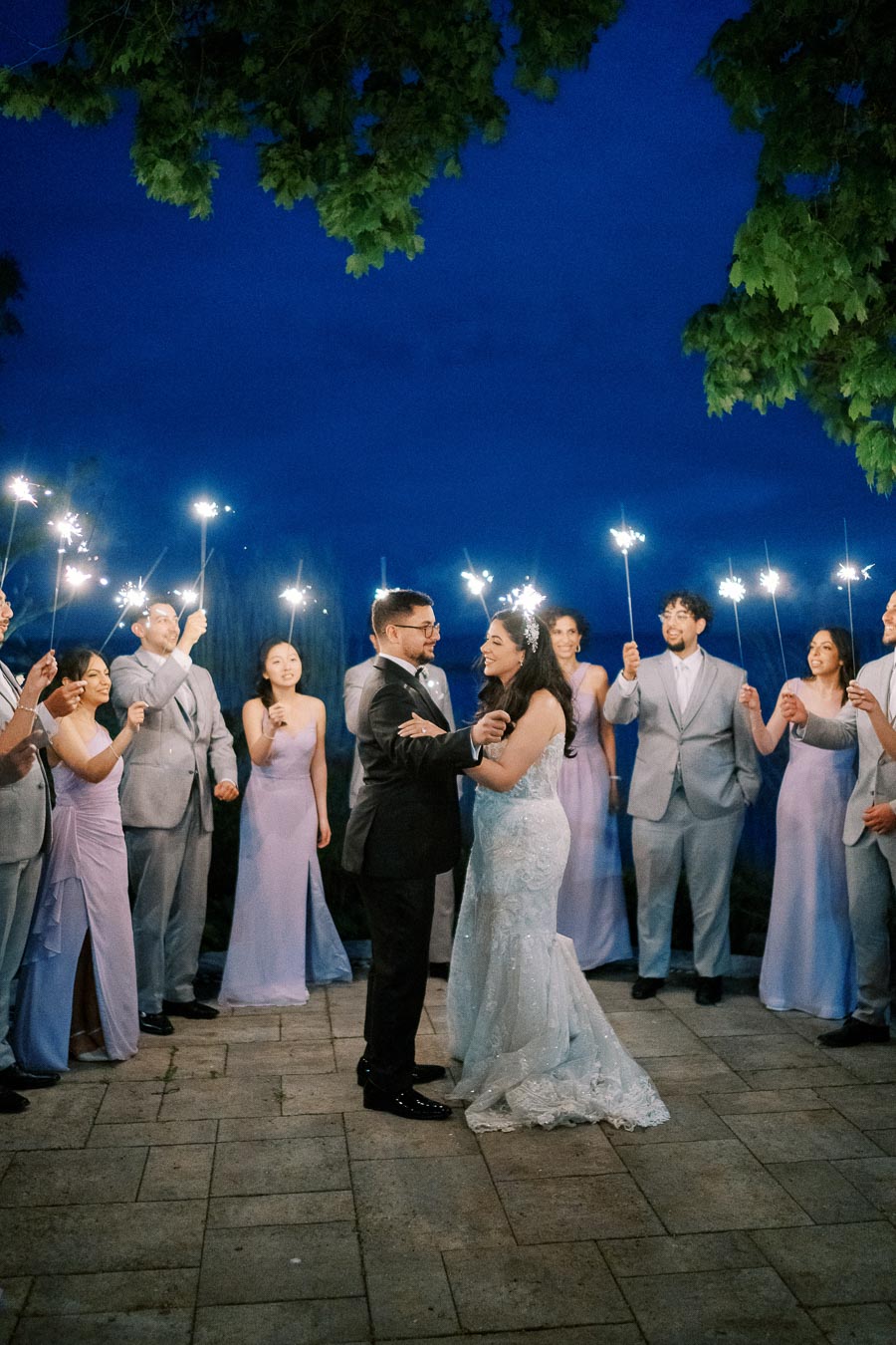 A bride and groom share a romantic first dance surrounded by a wedding party holding sparklers, under a vibrant night sky.