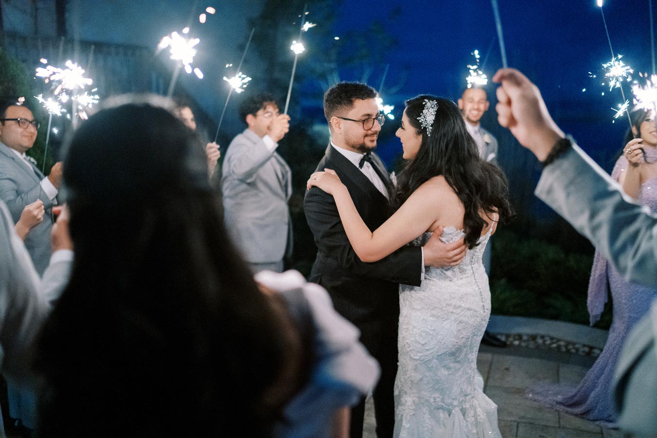 Newlywed couple shares a first dance surrounded by sparklers at an outdoor evening wedding celebration, with guests in formal attire creating a joyful ambiance.