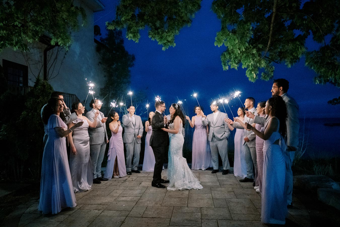 A wedding couple dances under a canopy of sparklers held by their guests in evening attire, surrounded by friends in lilac and gray outfits, set against a twilight outdoor backdrop.