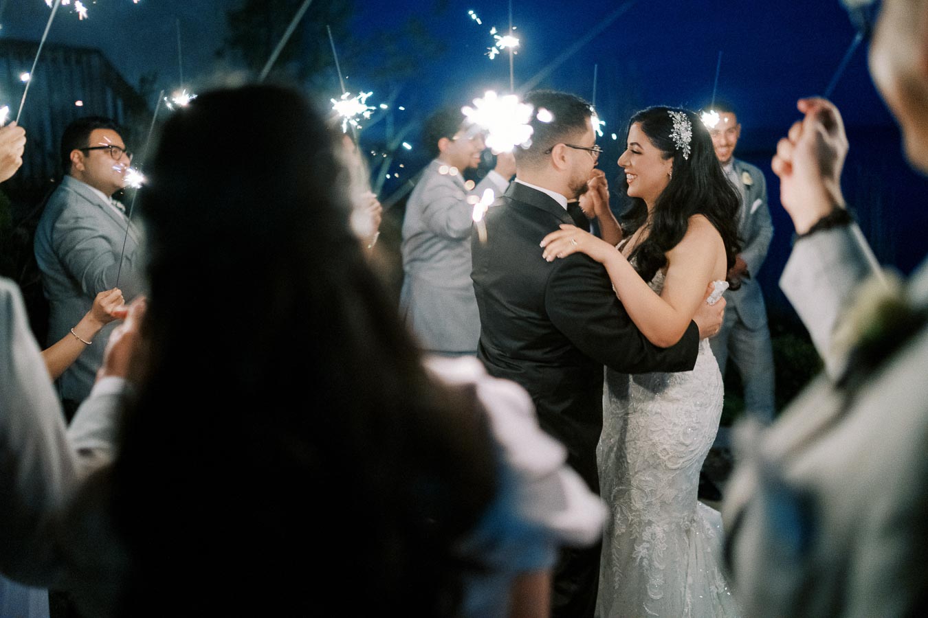 A bride and groom share a romantic dance surrounded by wedding guests holding sparklers during an evening celebration. The couple looks joyful, creating a magical and intimate atmosphere on their special day.