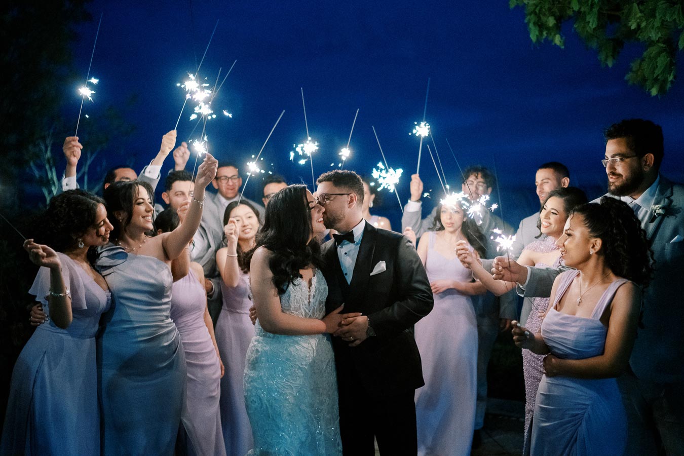 A newlywed couple shares a romantic moment surrounded by their wedding party holding sparklers against a vibrant night sky.