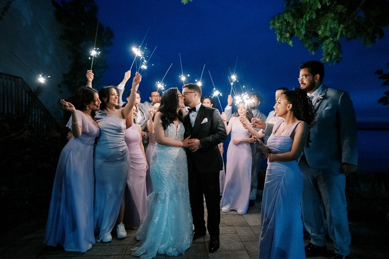 A newlywed couple embraces surrounded by a joyful wedding party holding sparklers at night, with the bride in a white lace gown and the groom in a black tuxedo, creating a magical evening celebration atmosphere.