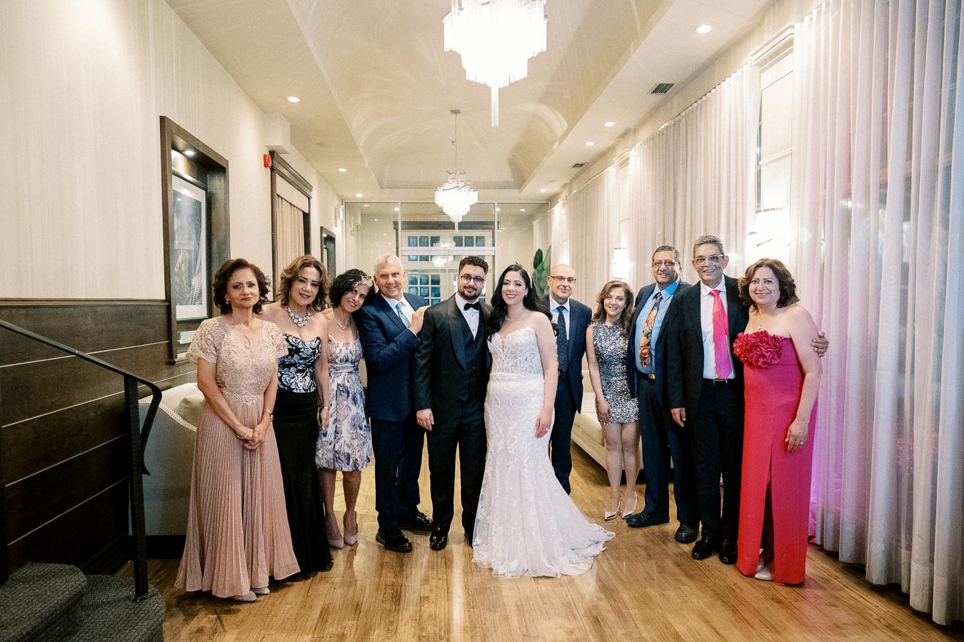 A joyful group of elegantly dressed people posing together at an indoor wedding event, with the bride and groom surrounded by family and friends, set in a beautifully lit venue with chandeliers.