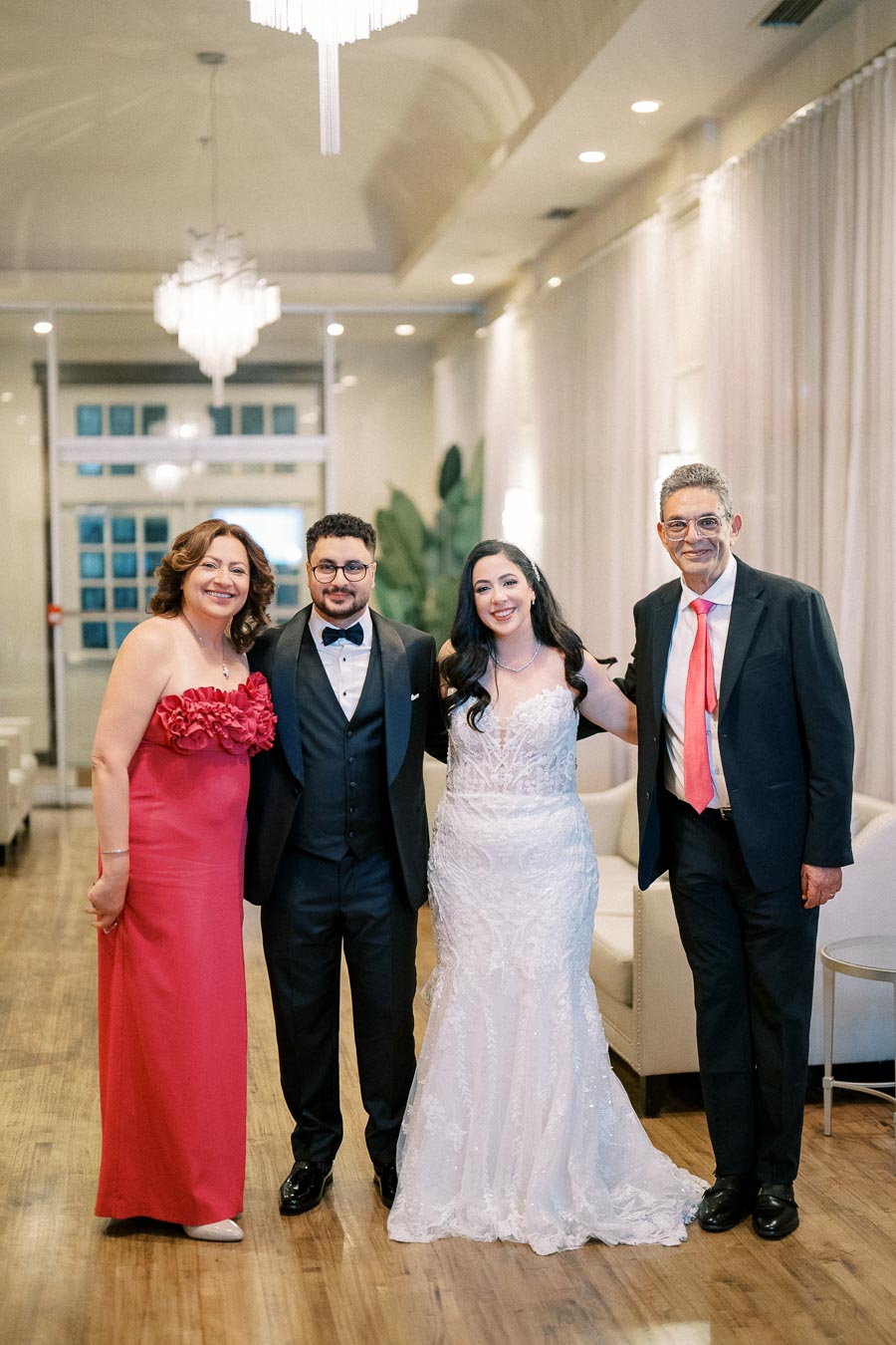A joyful wedding celebration with four elegantly dressed individuals posing in a beautifully decorated indoor venue, featuring chandeliers and luxurious furnishings.