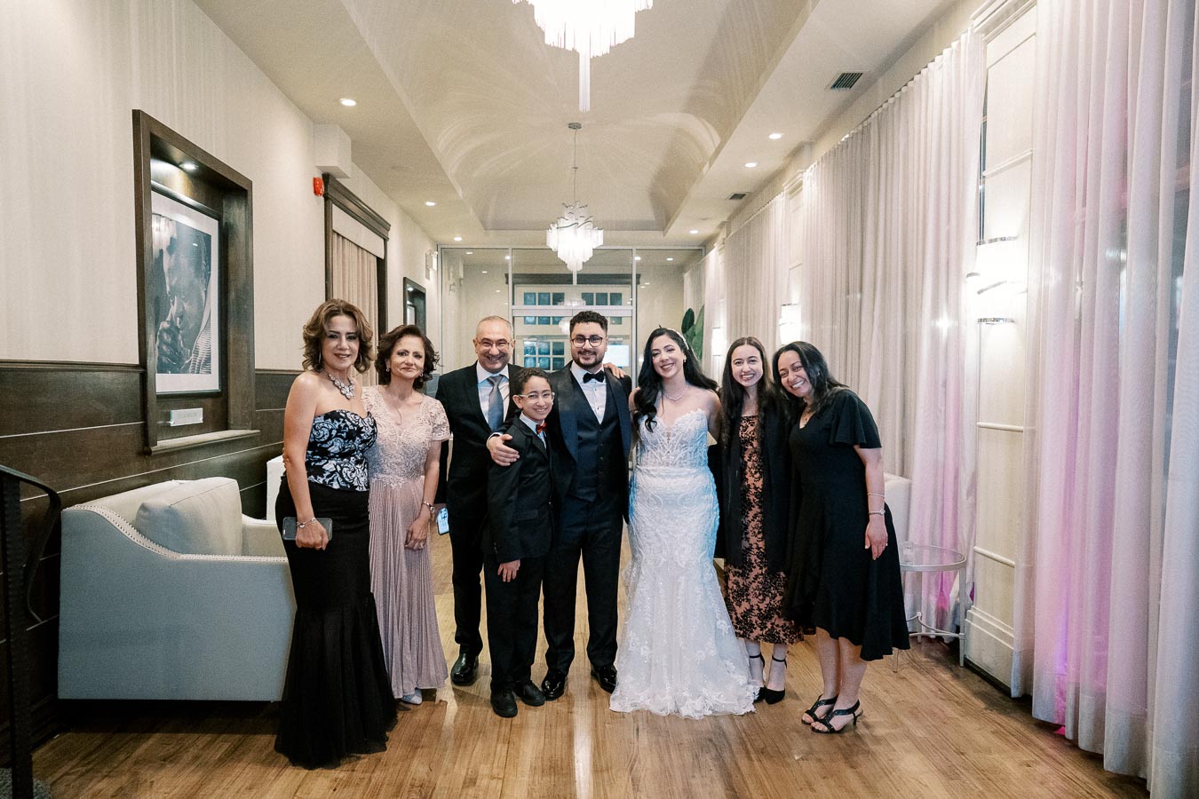 A happy group of people dressed elegantly at a wedding, standing together in a beautifully decorated indoor venue with chandeliers and soft lighting.
