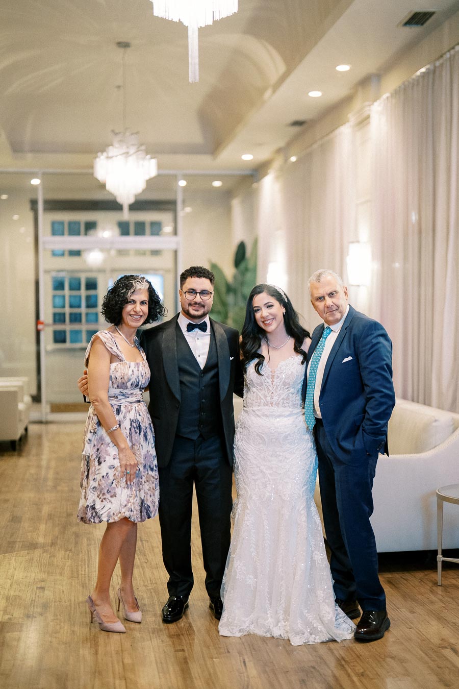 A wedding couple and two guests pose together in a beautifully decorated indoor venue, showcasing elegant attire and a joyful atmosphere.