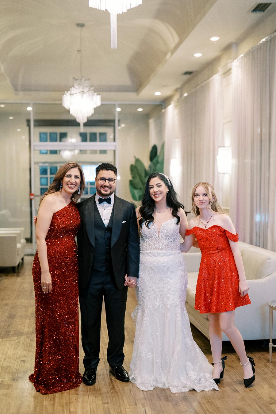 Group of elegantly dressed people smiling at a formal event, featuring a bride in a white lace gown, a groom in a tuxedo, and two women in sparkling red dresses, standing in a sophisticated, well-lit venue.