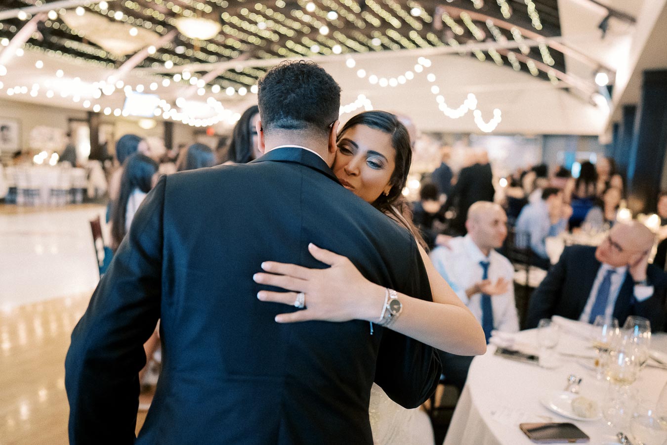 A person in formal attire embraces another at an elegant wedding reception, surrounded by warm lighting and guests seated at decorated tables.