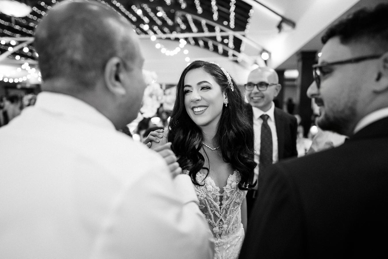 Black and white image of a joyful bride wearing an elegant lace wedding dress, surrounded by guests at a wedding reception with twinkling fairy lights in the background.