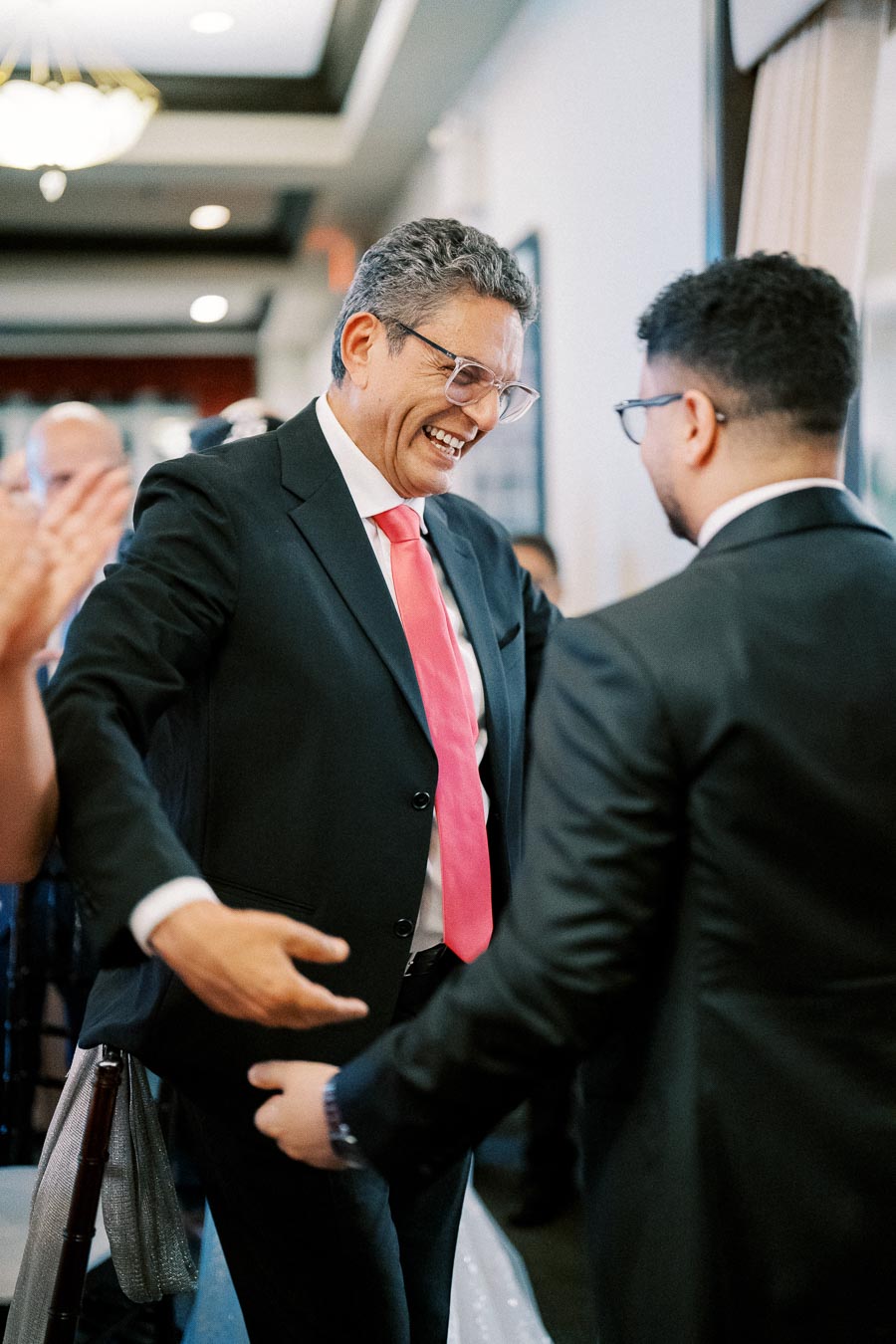 Two men in suits smiling and shaking hands at a formal event, one wearing a bright pink tie and glasses, capturing a moment of congratulation and celebration.
