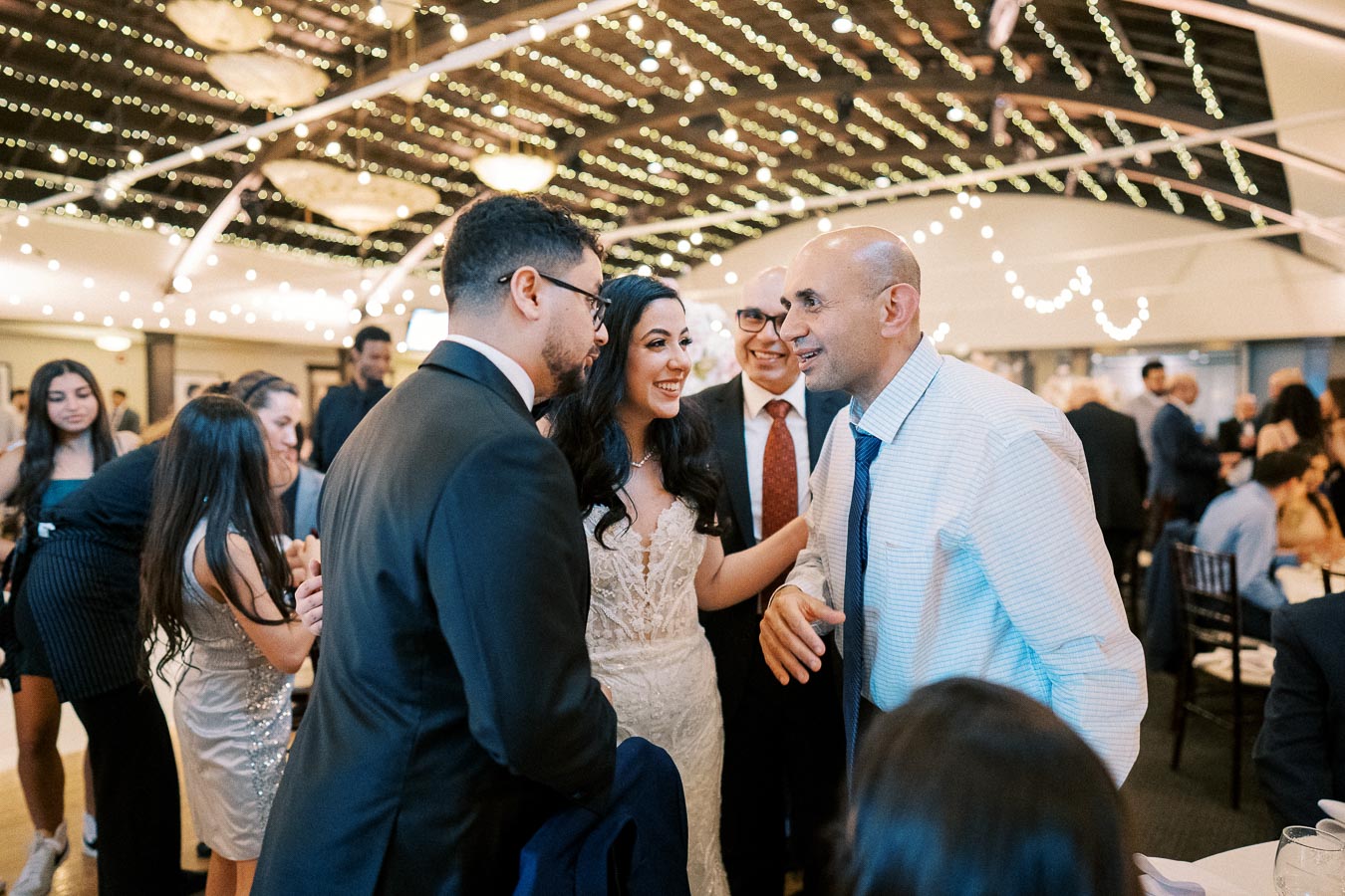 A joyful scene at a wedding reception with guests socializing under decorative string lights in an elegant venue.