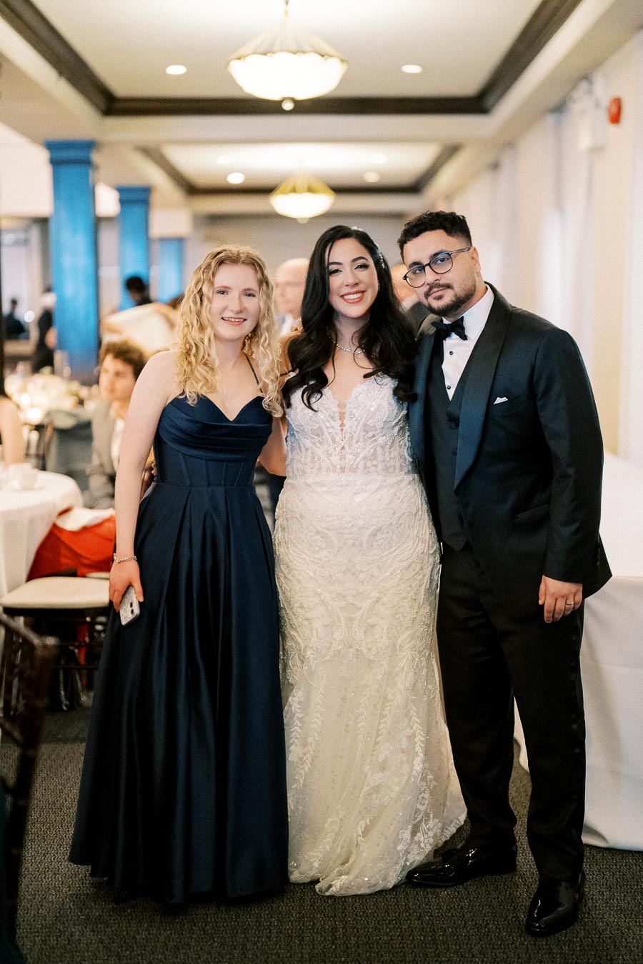 A bride in a lace wedding gown stands between a man in a black tuxedo and a woman in a navy blue dress, smiling at a wedding reception in a brightly lit banquet hall.