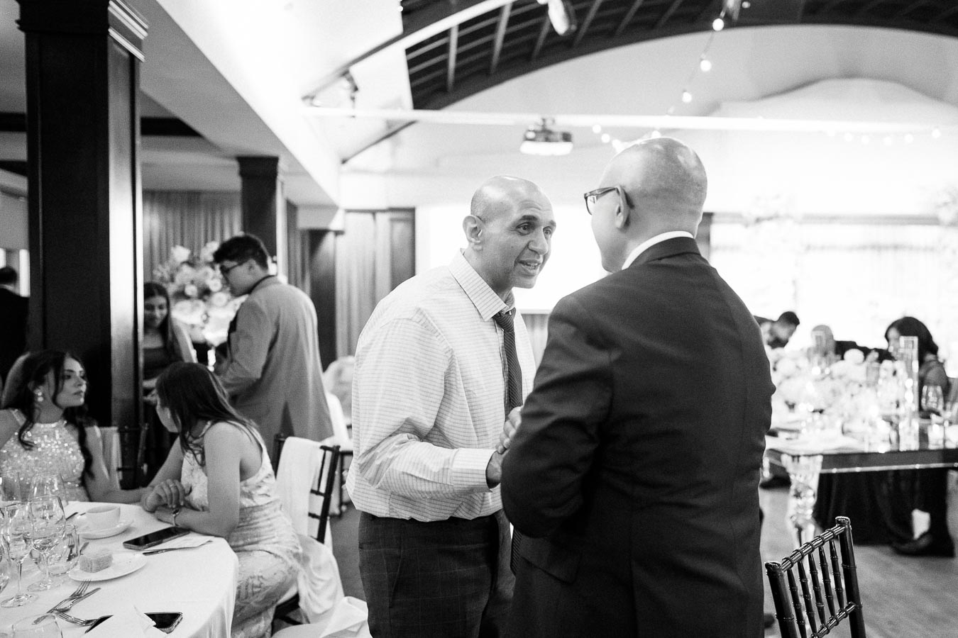 Black and white image of two men in conversation at a formal event, surrounded by seated guests at elegantly set tables.